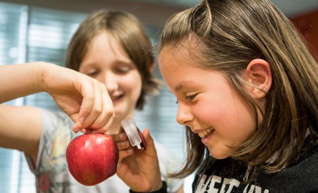 Due bambini osservano con una lente di ingrandimento una mela Alto Adige IGP e si divertono.